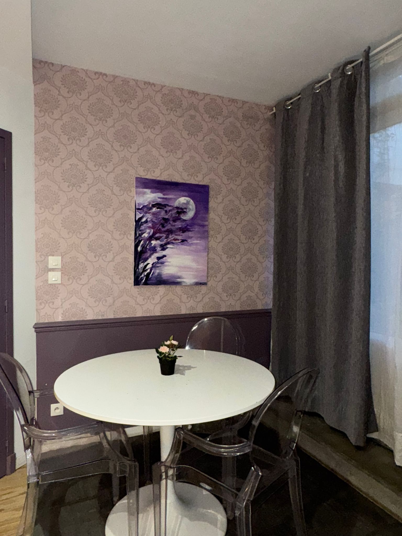 Een eetgedeelte met een ronde witte tafel, vier transparante stoelen, een kleine potplant en een aan de muur hangend schilderij van een paars landschap met een maan.