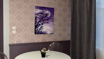Een eetgedeelte met een ronde witte tafel, vier transparante stoelen, een kleine potplant en een aan de muur hangend schilderij van een paars landschap met een maan.