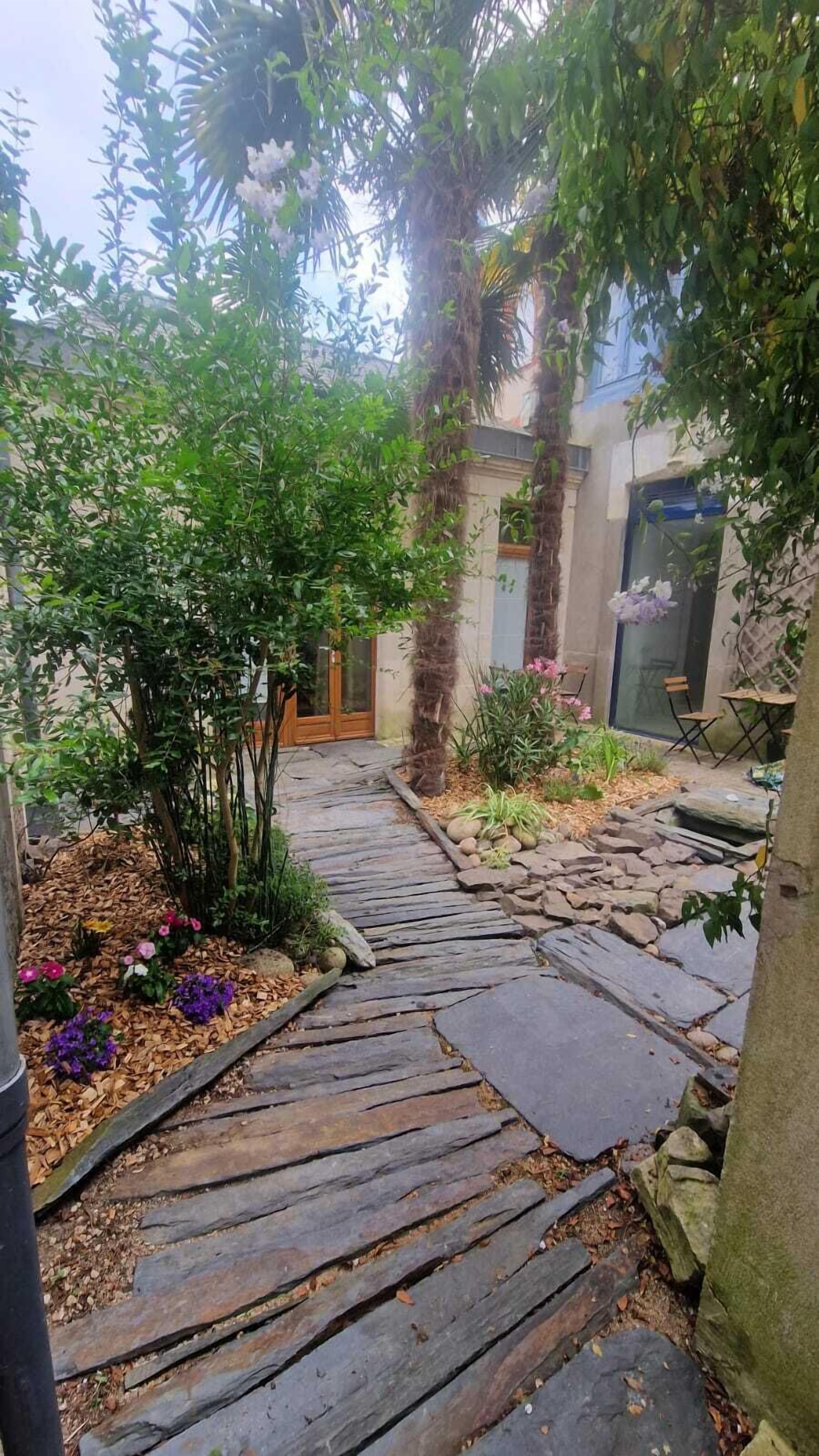 A stone pathway leading to a wooden door, surrounded by lush greenery and flowering plants.