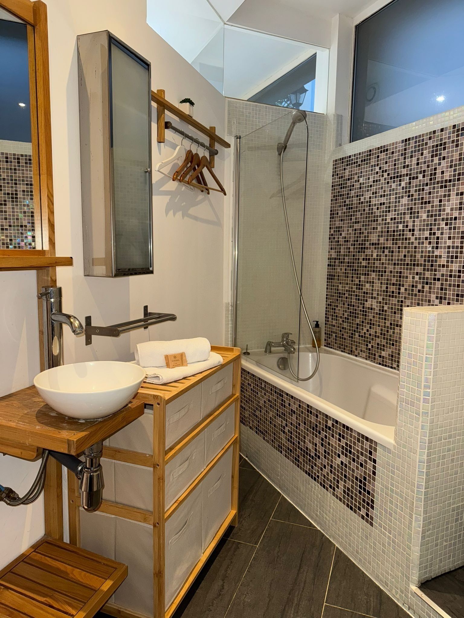 Et moderne badeværelse med et stort spejl, en trævasketøjsbord, en hvid vask, et badekar med mosaikfliser og et bruseområde.