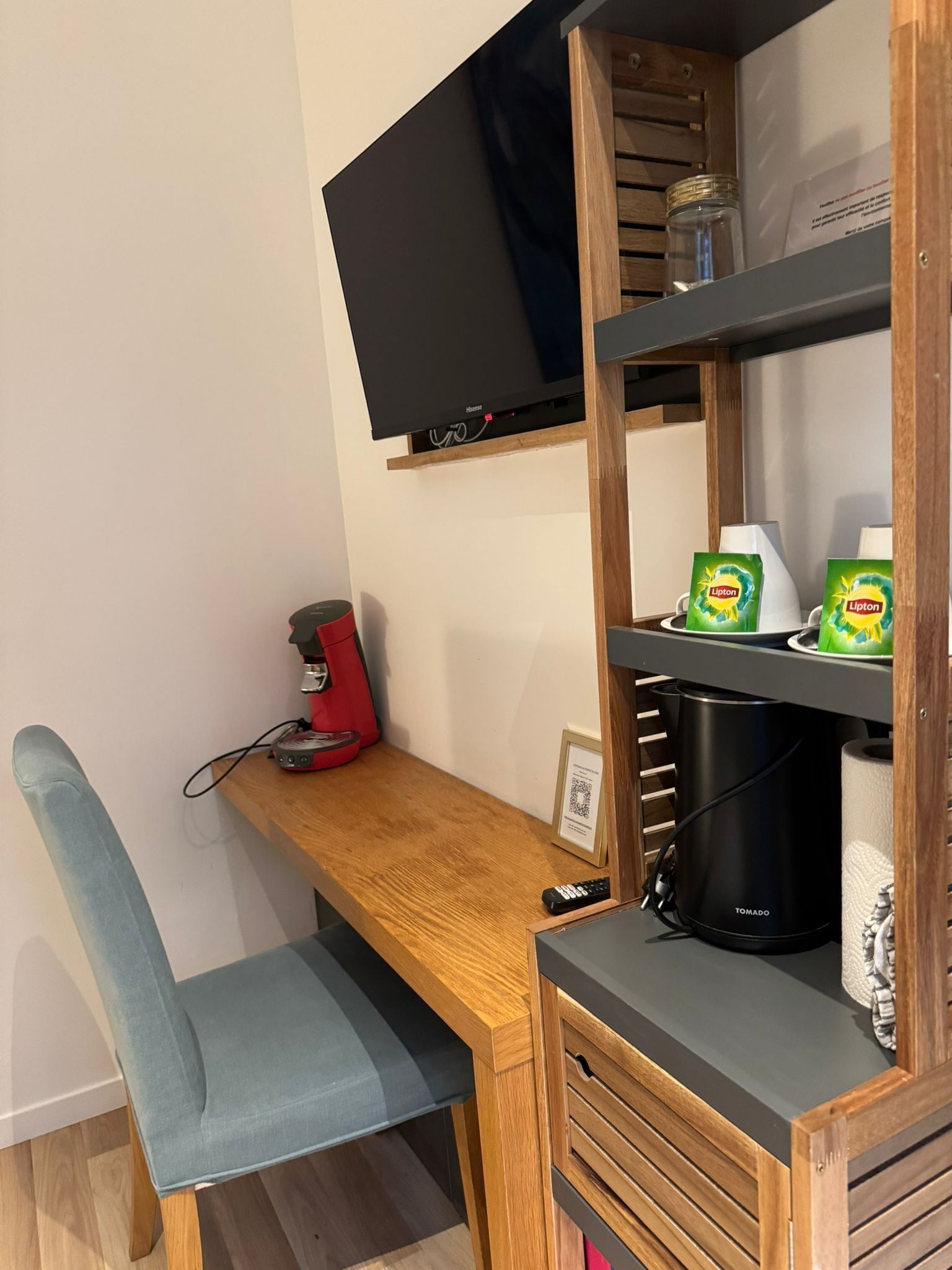 Quarto pequeno com escrivaninha de madeira, uma cadeira, televisão fixada na parede, cafeteira, chaleira e uma prateleira com caixas de chá Lipton.