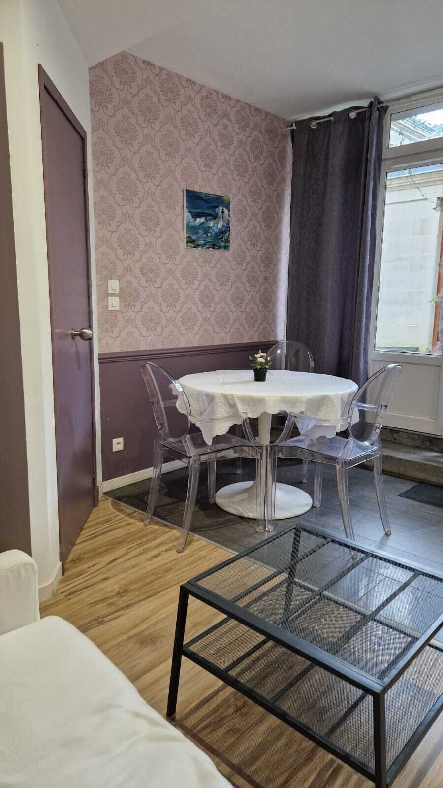 Et værelse med et rundt bord, transparente stole, en lille potteplante og et maleri på væggen.