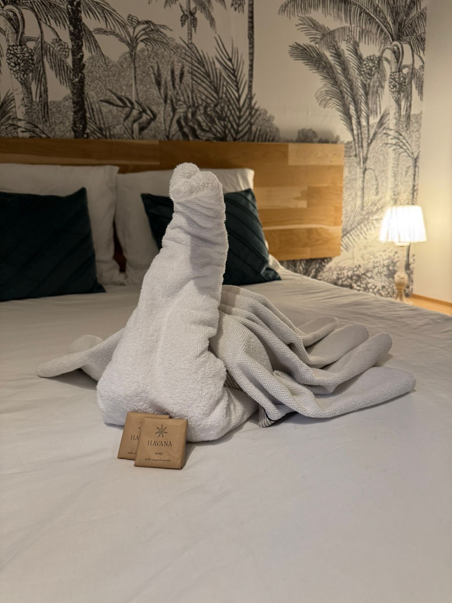 Ein ordentlich bezogenes Bett mit einem weißen Handtuch, einem Holzschild und einer Lampe auf einem Nachttisch.
