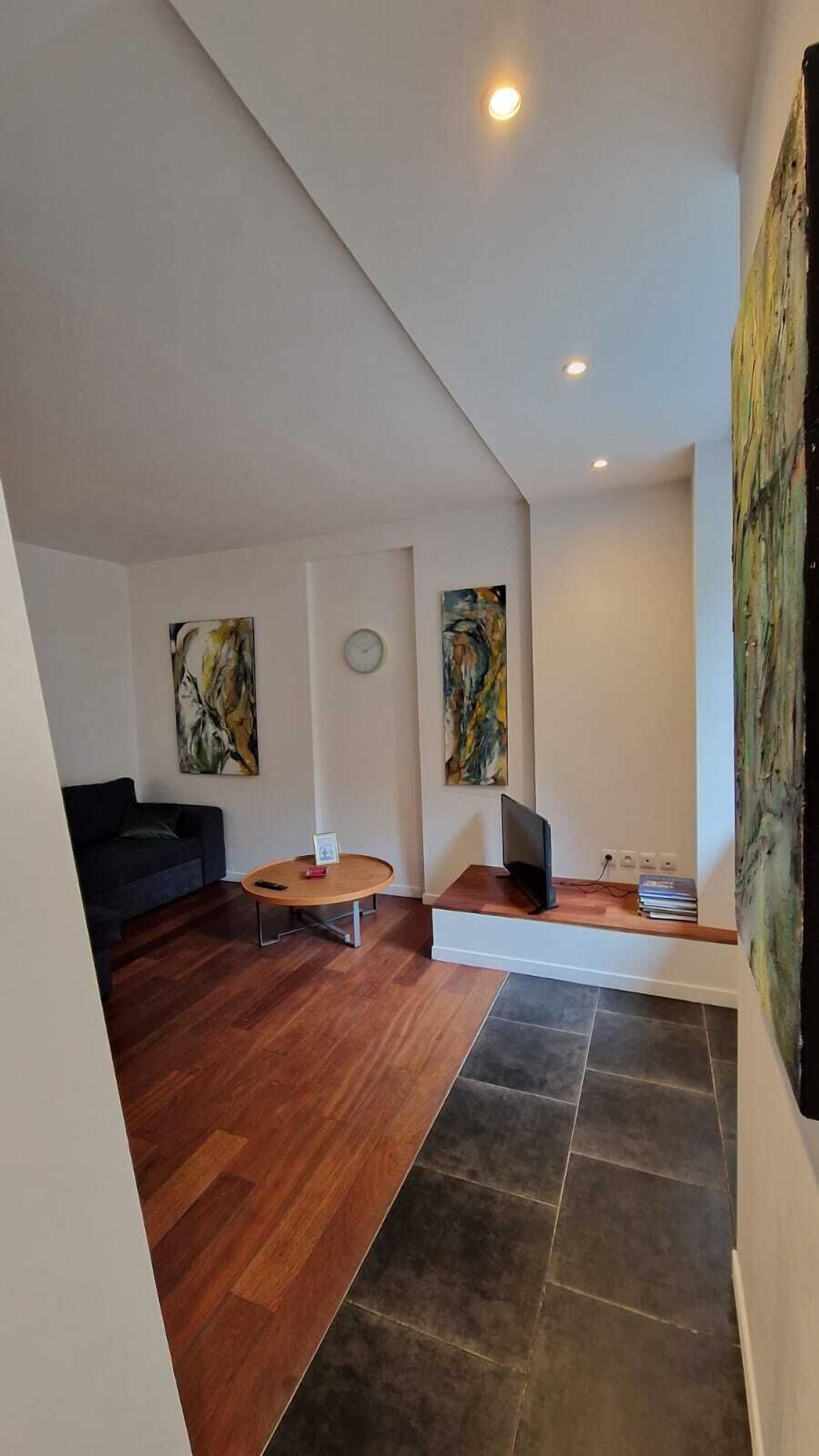 Un salon moderne avec un canapé de couleur sombre, une table basse ronde et un téléviseur à écran plat posé sur un meuble. Les murs sont ornés de tableaux abstraits et une horloge est accrochée au-dessus de la table basse.
