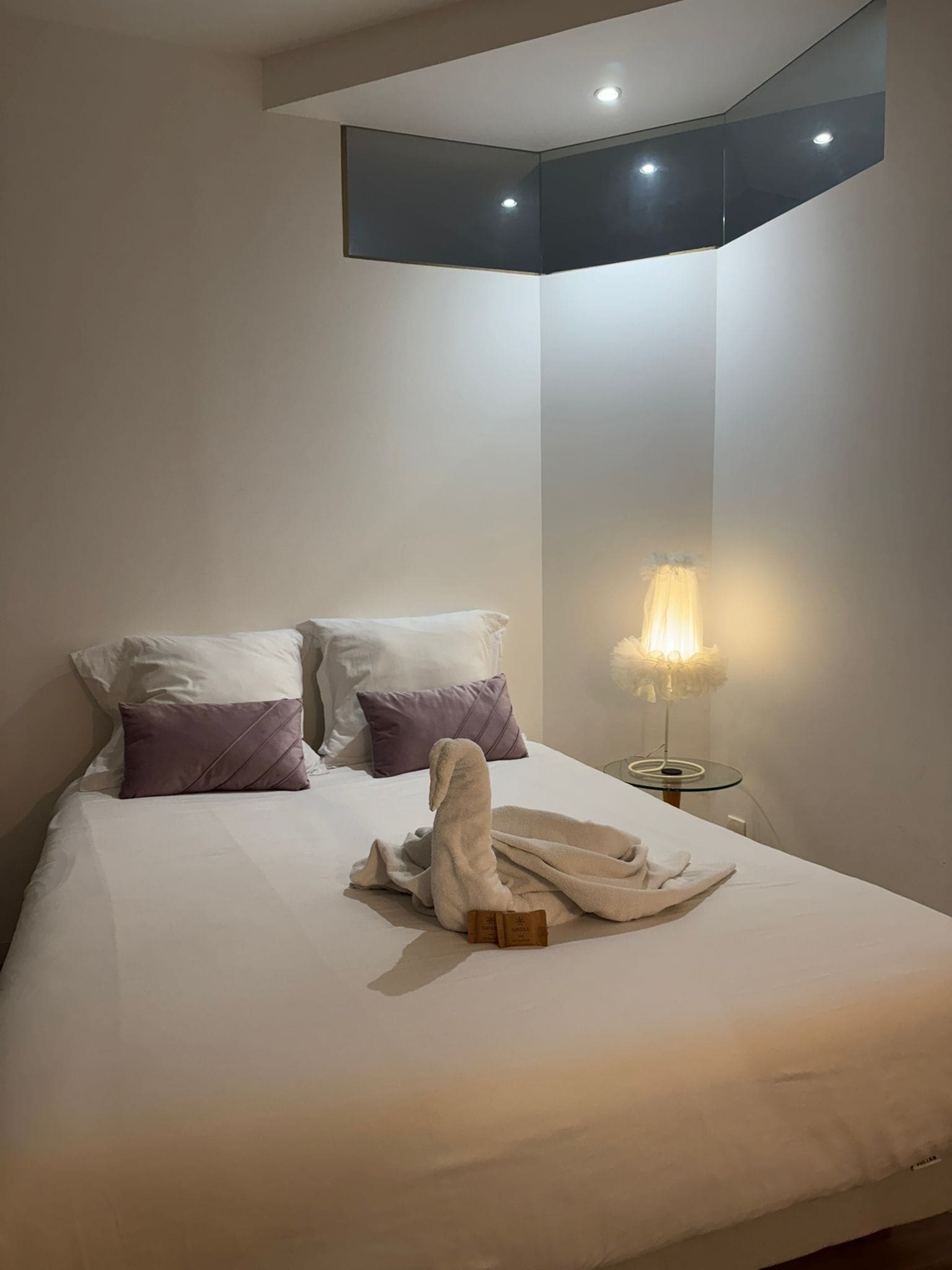 Una cama bien hecha con almohadas blancas y moradas, una bata blanca y una mesita de noche con una lámpara.