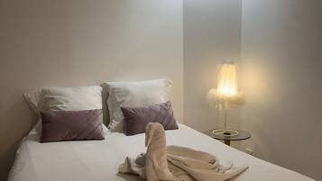 Una cama bien hecha con almohadas blancas y moradas, una bata blanca y una mesita de noche con una lámpara.