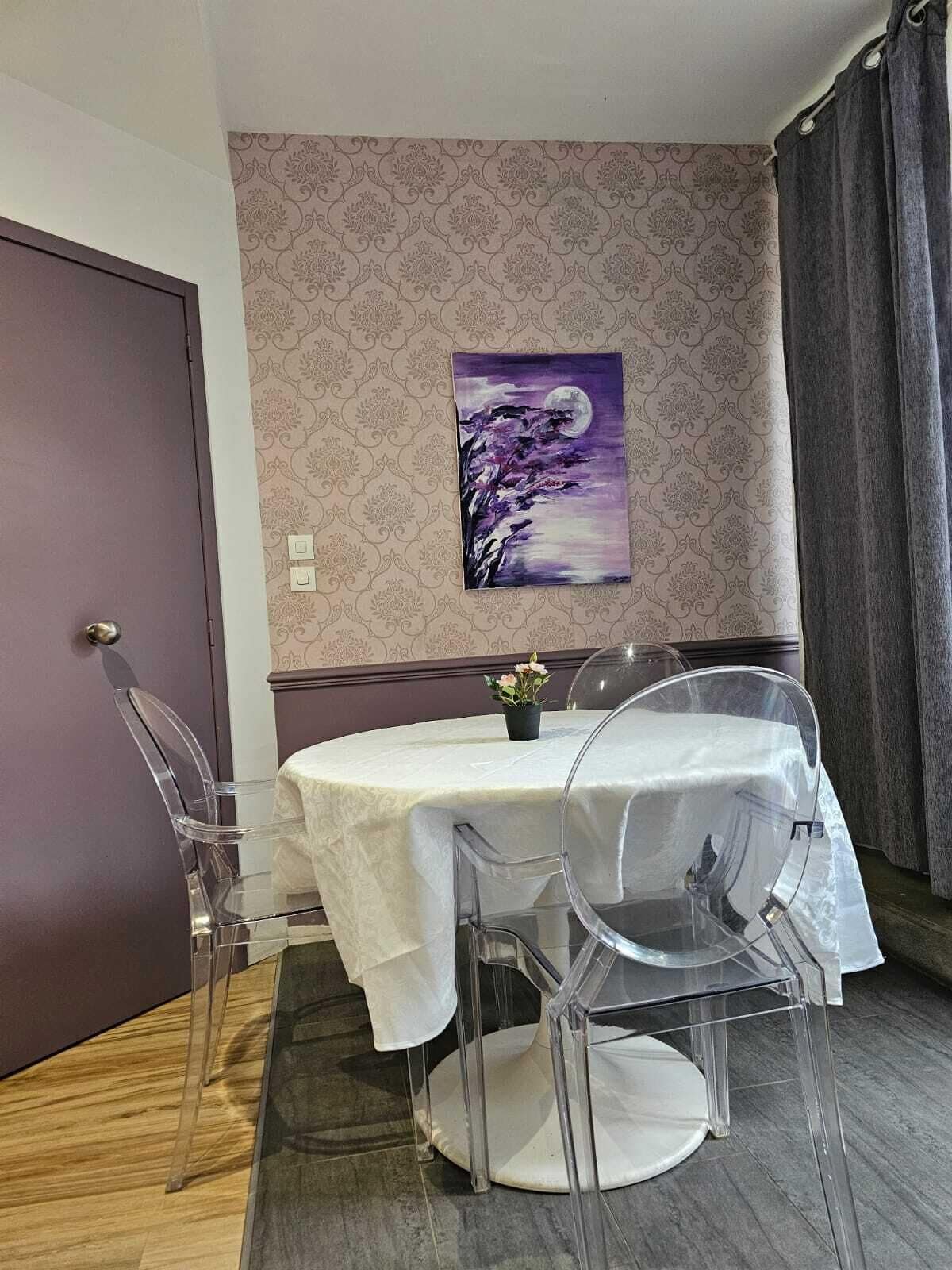 Uma sala de jantar com uma mesa redonda, cadeiras transparentes, toalha de mesa branca, um quadro roxo na parede e uma porta.
