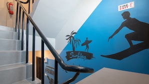 A staircase with a blue wall featuring a surfing theme, a fire exit sign, and a fire extinguisher. - Hip Hostel (Patong)