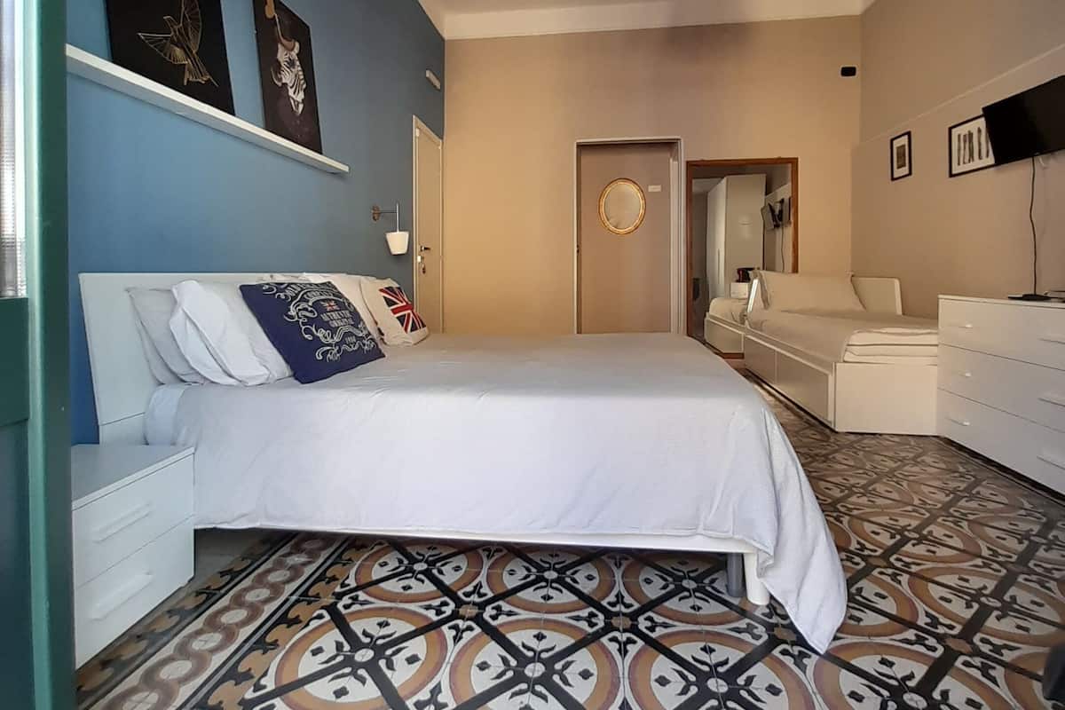 Un dormitorio con piso estampado, una cama con ropa de cama blanca, un aparador, una silla y una lámpara en el techo.