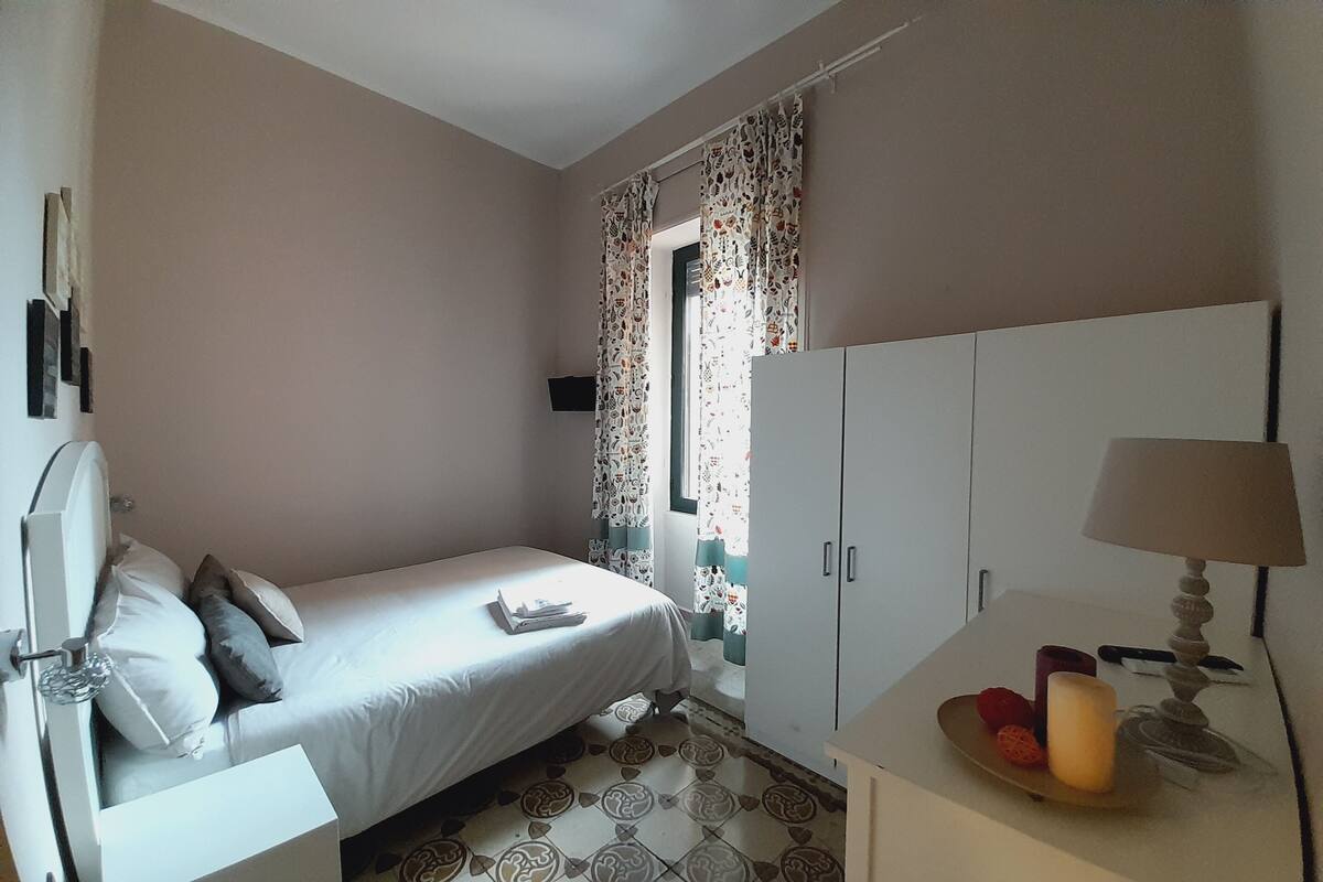 Un dormitorio con cama, armario, mesita de noche con lámpara y ventana con cortinas.
