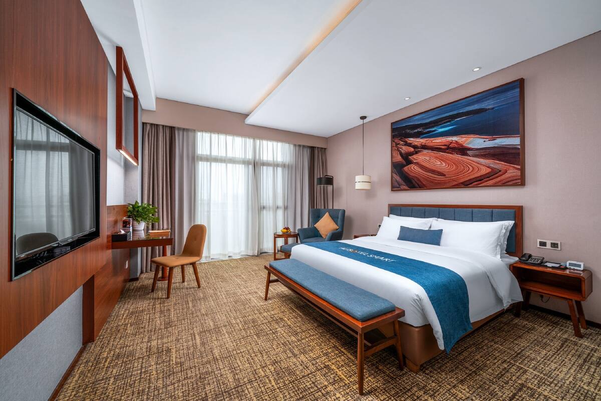 A modern hotel room with a large bed, a desk with a TV, a bench, and a painting on the wall.