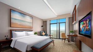 A hotel room with a large bed, a desk, a chair, a TV, and a large window with a view of the sky.