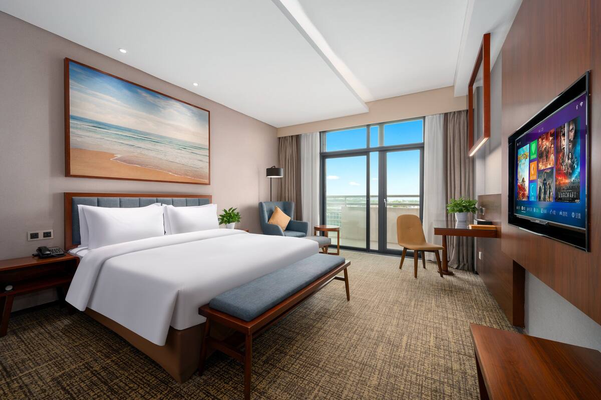A hotel room with a large bed, a desk, a chair, a TV, and a large window with a view of the sky.