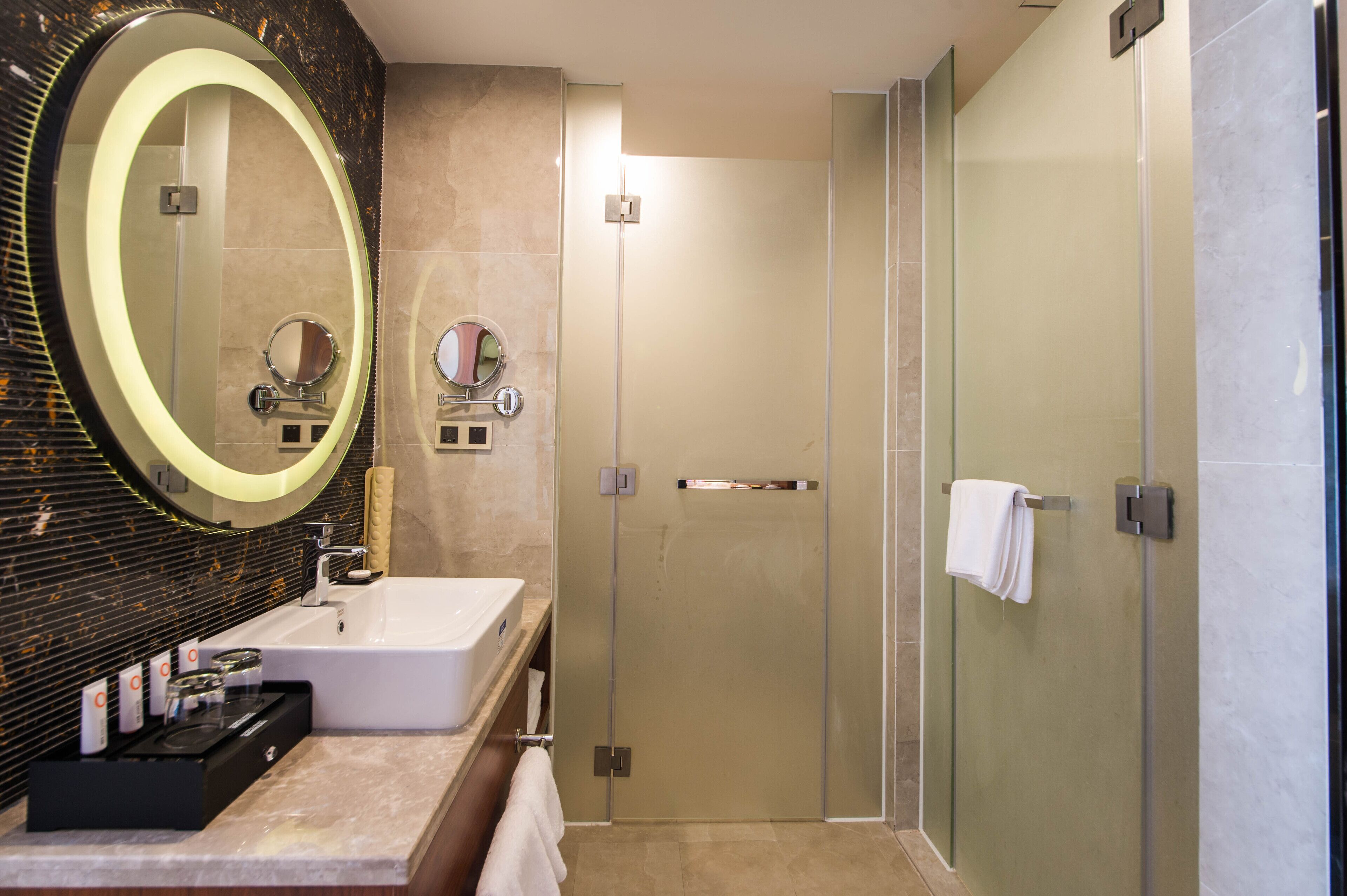 A modern bathroom with a large oval mirror, a white sink, a glass shower enclosure, and a towel rack.