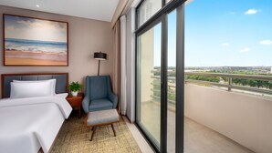 A hotel room with a bed, a chair, a nightstand with a lamp, a painting of a beach, and a balcony with a city view.