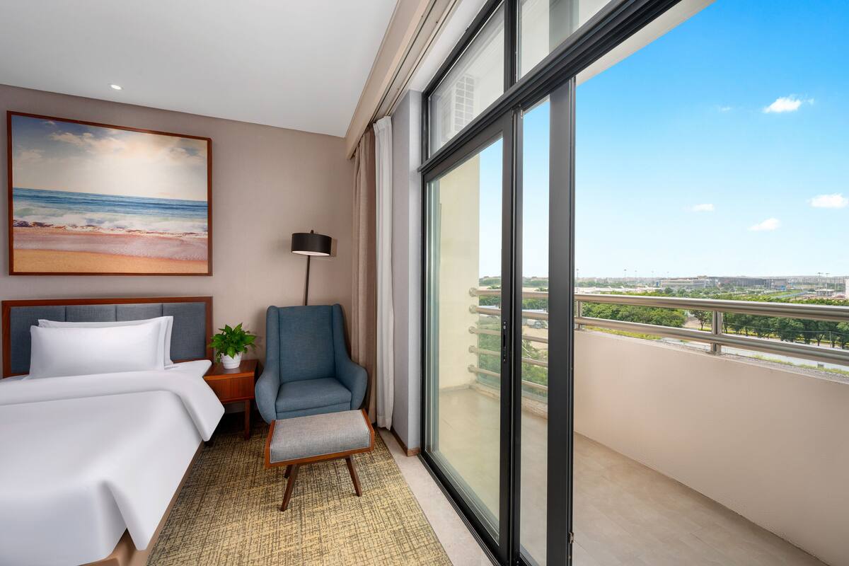 A hotel room with a bed, a chair, a nightstand with a lamp, a painting of a beach, and a balcony with a city view.