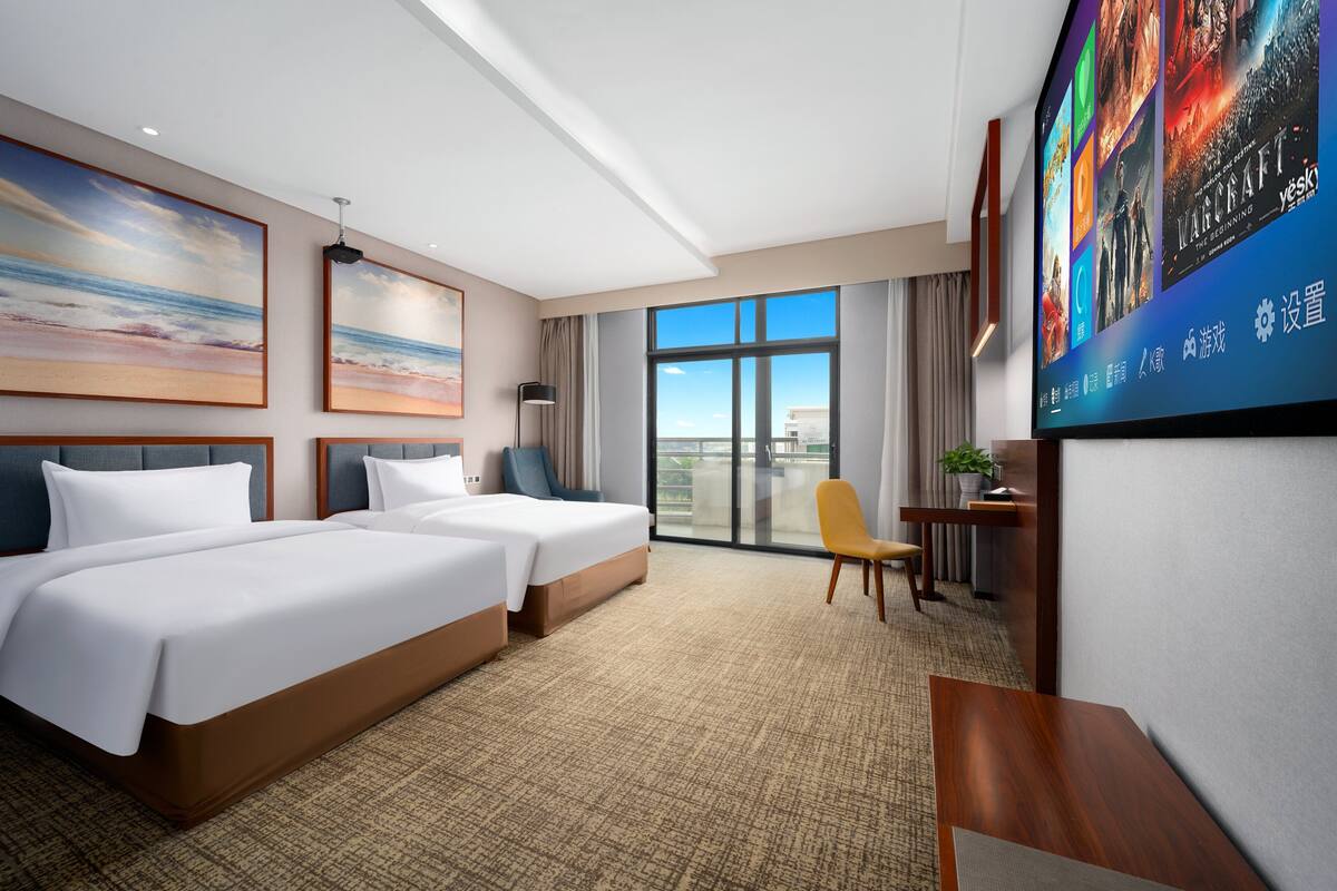 A hotel room with two beds, a large window, a TV screen, and a desk.