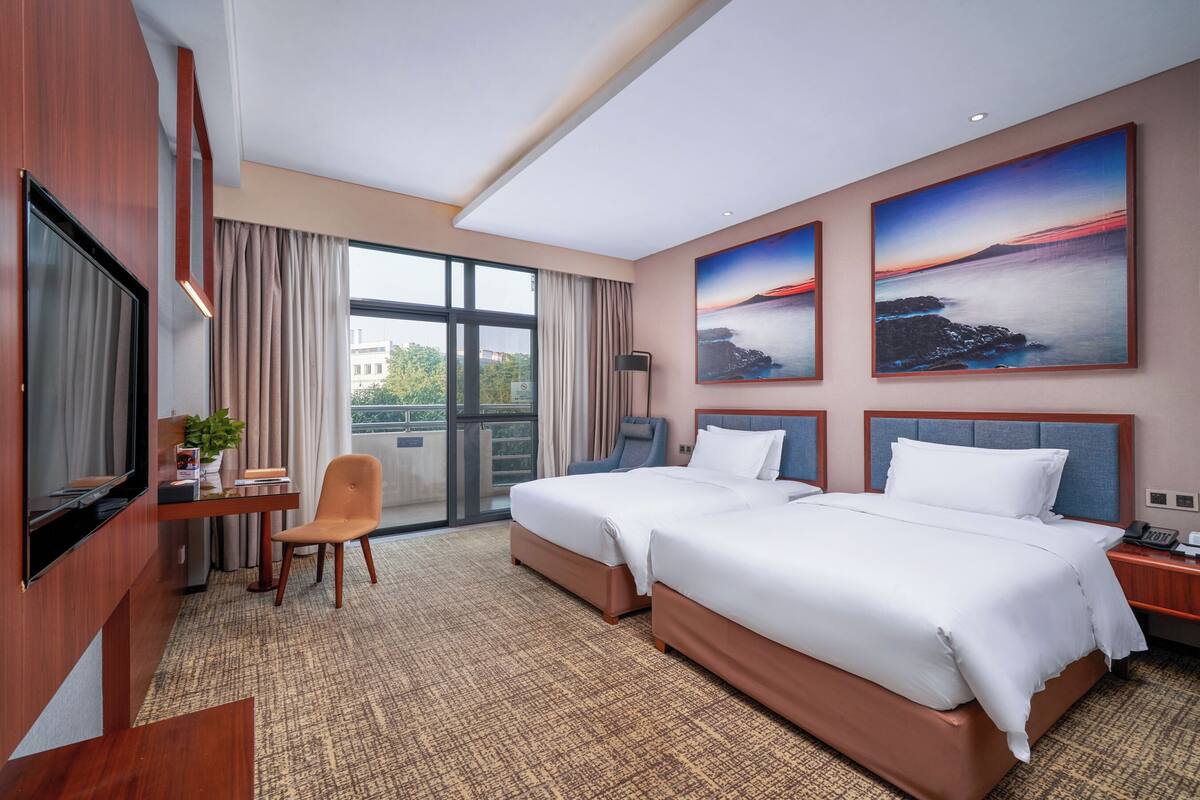 A hotel room with two beds, a desk with a chair, a TV, and large windows with curtains.