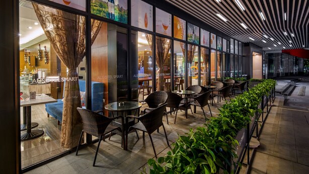 A modern hotel with a long corridor lined with tables and chairs, large windows, and a patterned ceiling.