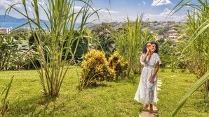 A woman in a white dress standing in a lush garden with tall grasses and a view of a coastal town in the background. - Créole Bay - Suites and Spa (Fort-de-France)