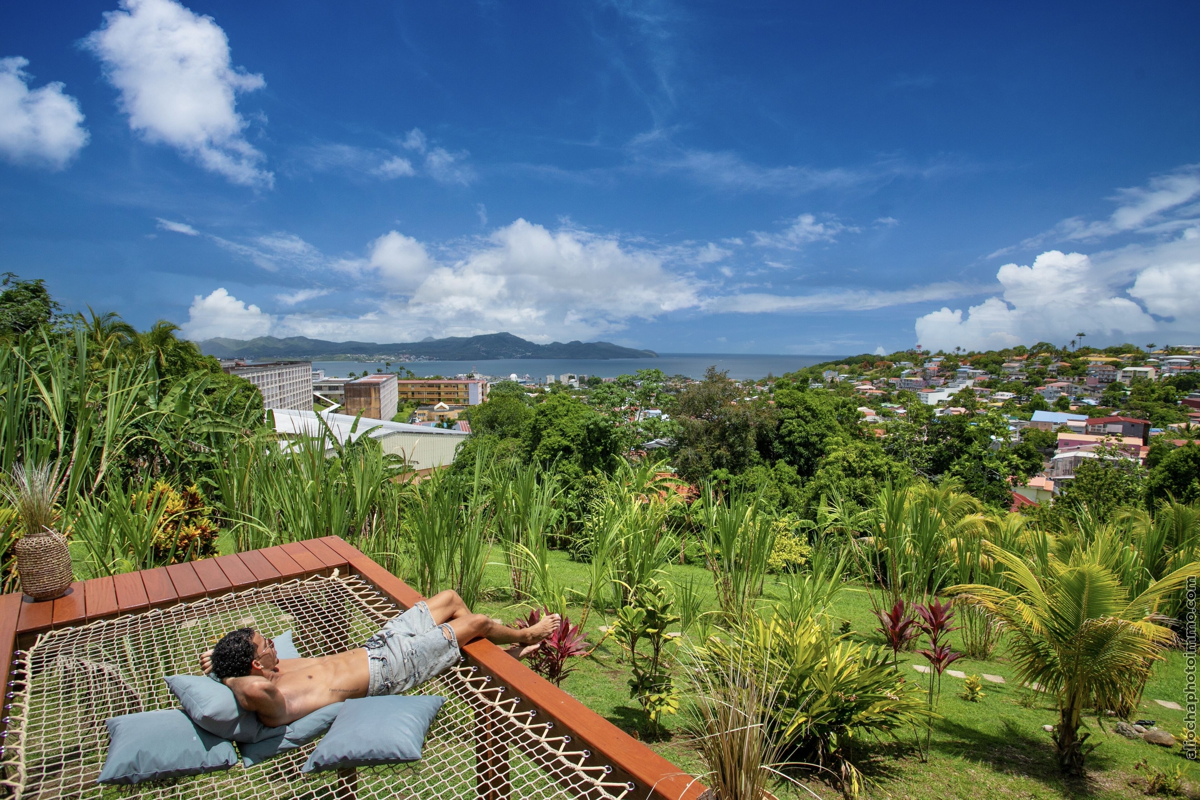 A hammock on a wooden deck overlooking a lush garden and a coastal town.