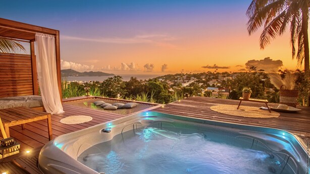 A hot tub on a wooden deck with a view of a cityscape and mountains at sunset. - Créole Bay - Suites and Spa (Fort-de-France)