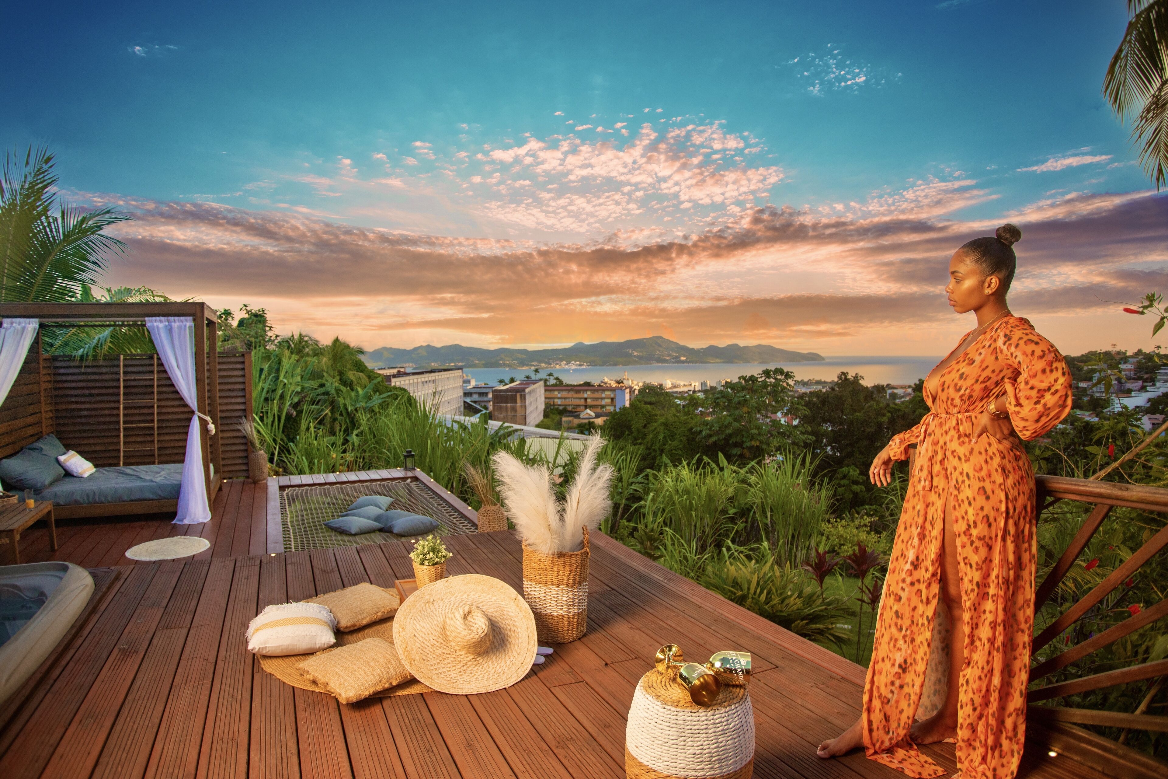 Une terrasse en bois offrant une vue sur l’océan et les montagnes, un coin salon avec un canapé et des coussins, ainsi que des objets décoratifs comme un chapeau et des vases.