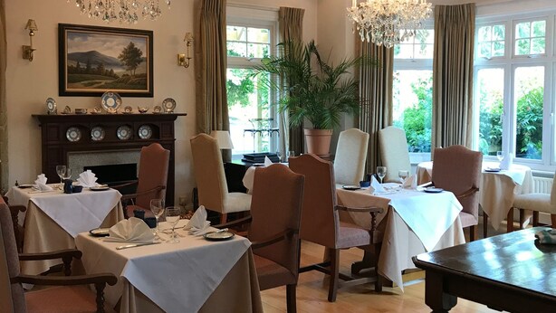A dining area with wooden tables, chairs, chandeliers, a fireplace, and a large window with curtains. - The Pines Hotel (Slough)