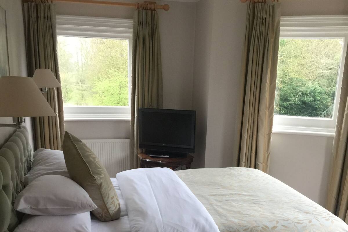 A hotel room with a large bed, a television on a wooden stand, and windows with curtains.