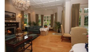 A spacious living room with a fireplace, comfortable seating, and large windows offering a view of greenery. - The Pines Hotel (Slough)