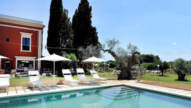 A swimming pool with white lounge chairs and umbrellas, surrounded by a red building and tall trees. - Agriturismo Santa Chiara (Alezio)