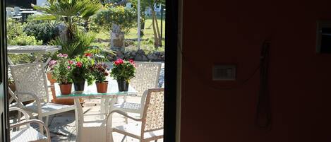 Un patio al aire libre con mobiliario de mimbre blanco, plantas en macetas y vistas a un jardín.