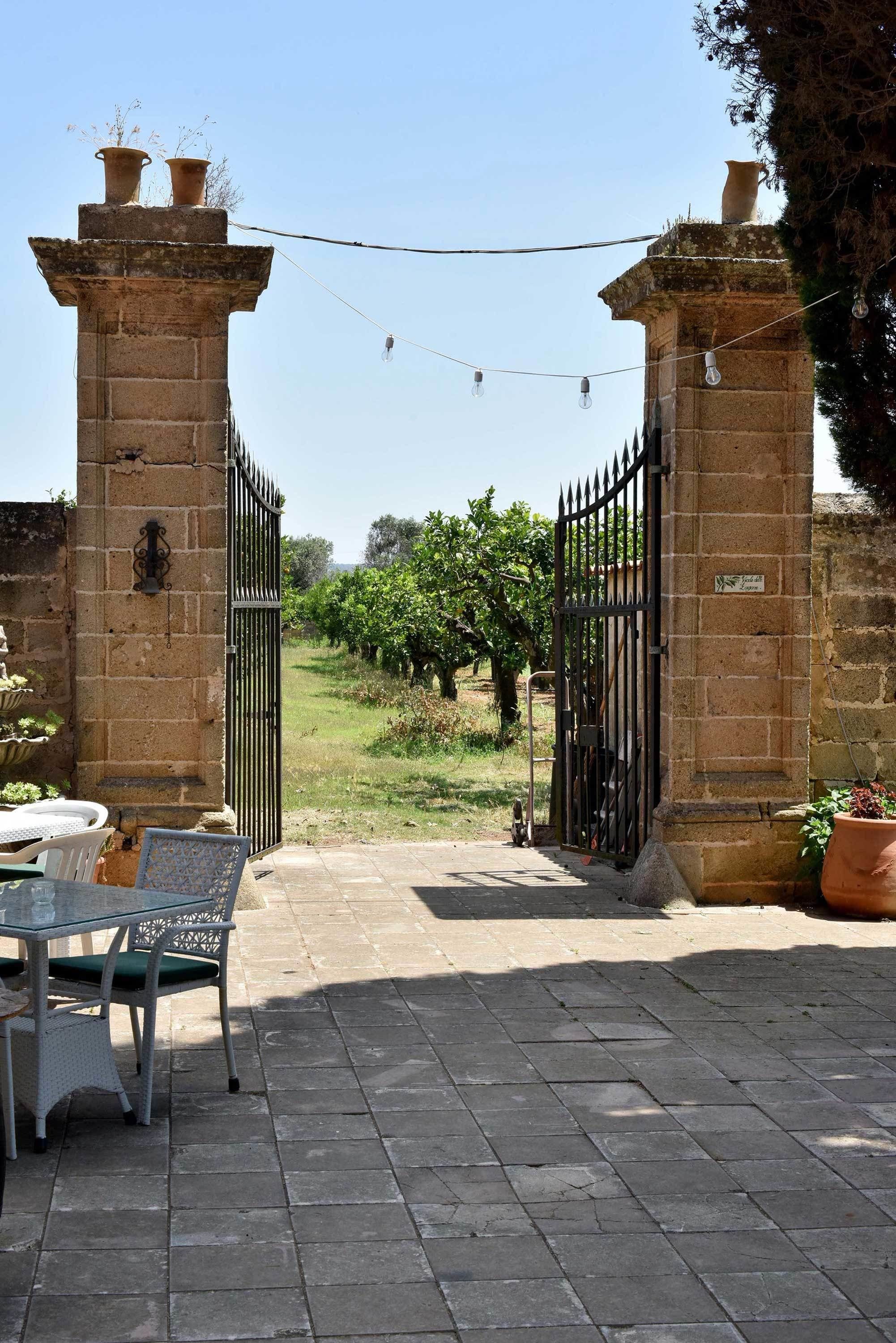 Une entrée fermée par un portail, avec une table et des chaises, un mur de pierre et une vue sur un vignoble.
