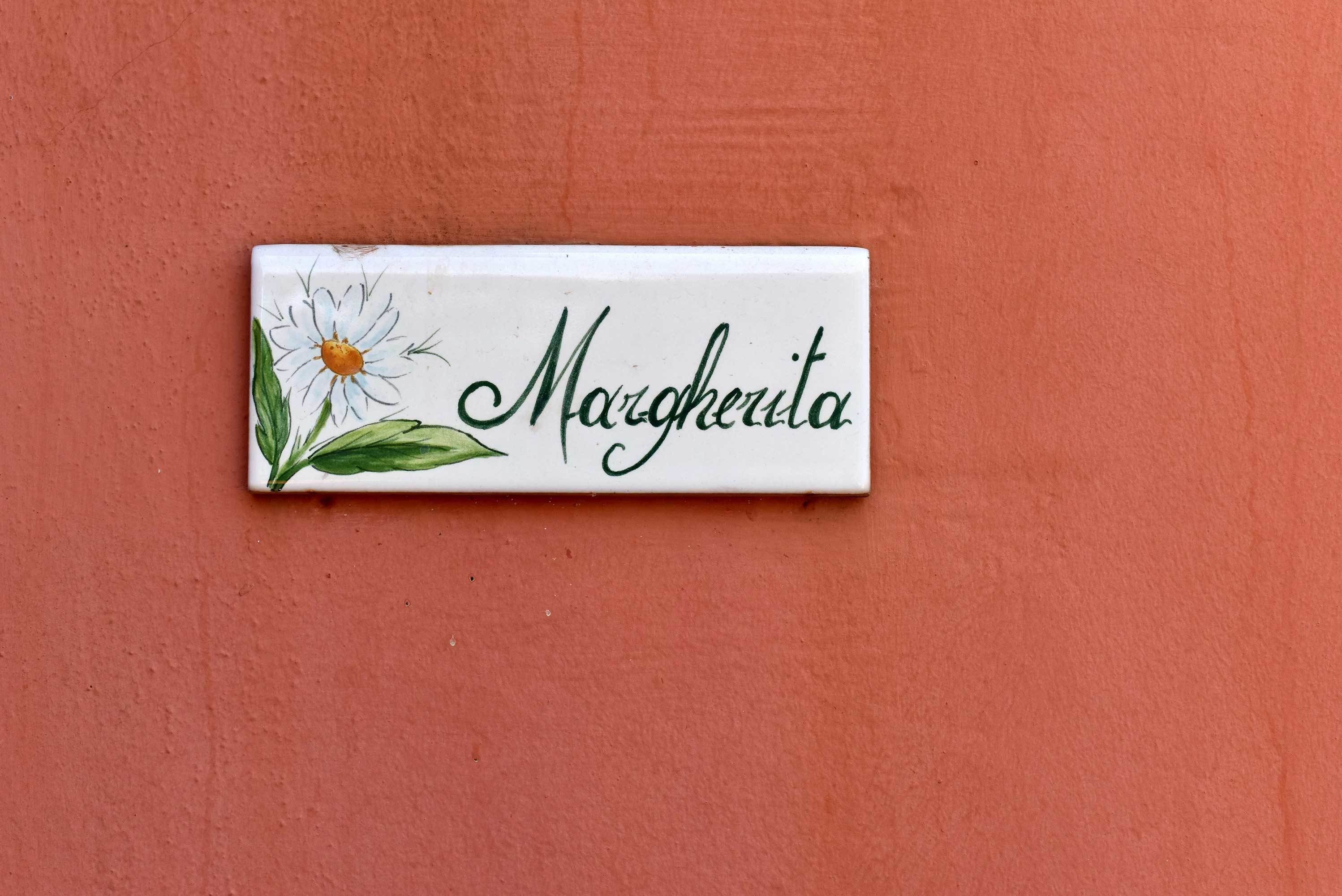 A white rectangular sign with the name "Margherita" in cursive, accompanied by a daisy illustration.
