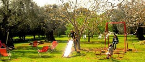 Um parque com parquinho, incluindo balanços e escorrega, cercado por árvores e bancos.