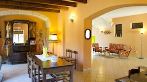 A dining area with a wooden table and chairs, a kitchen with a brick archway, and a living room with a sofa and chairs. - Agriturismo Santa Chiara (Alezio)