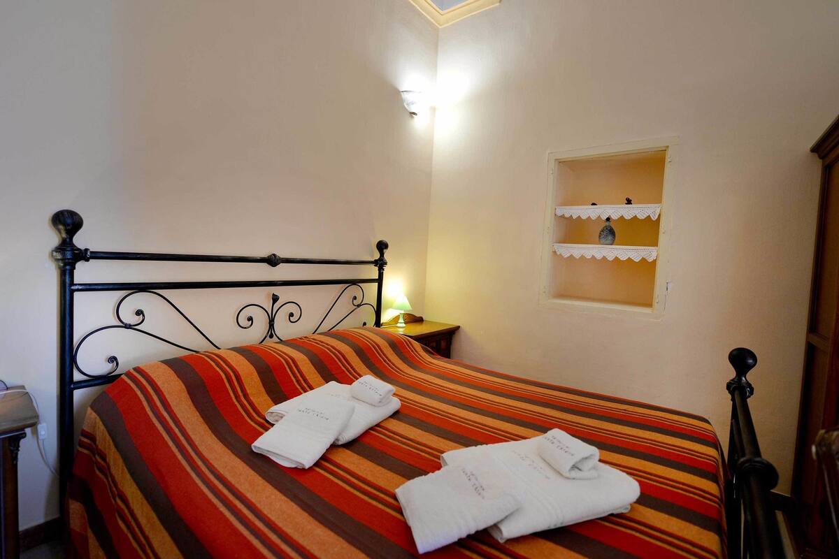 A neatly made bed with striped bedding and two towels, a bedside lamp, and a small shelf with decorative items.