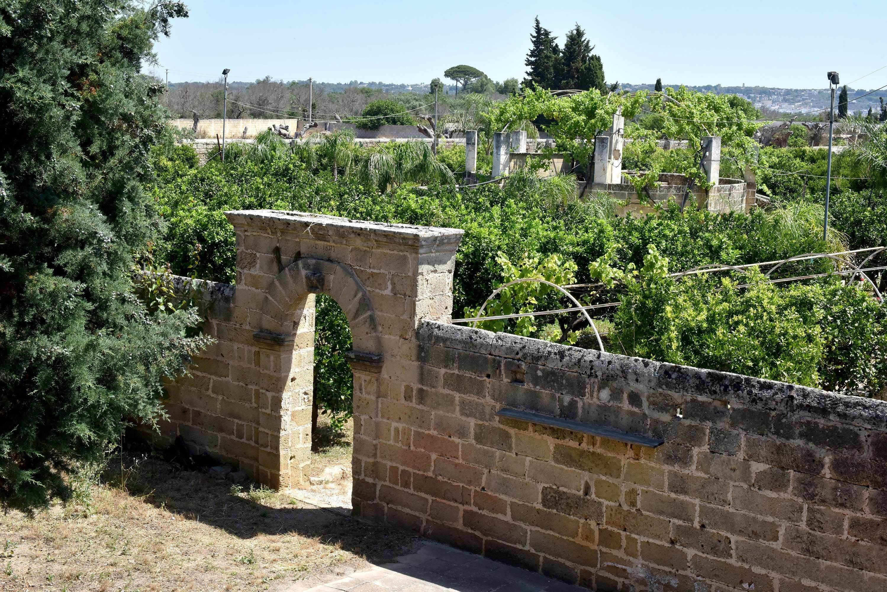 An arched stone structure in a garden with dense greenery and a distant view of buildings.