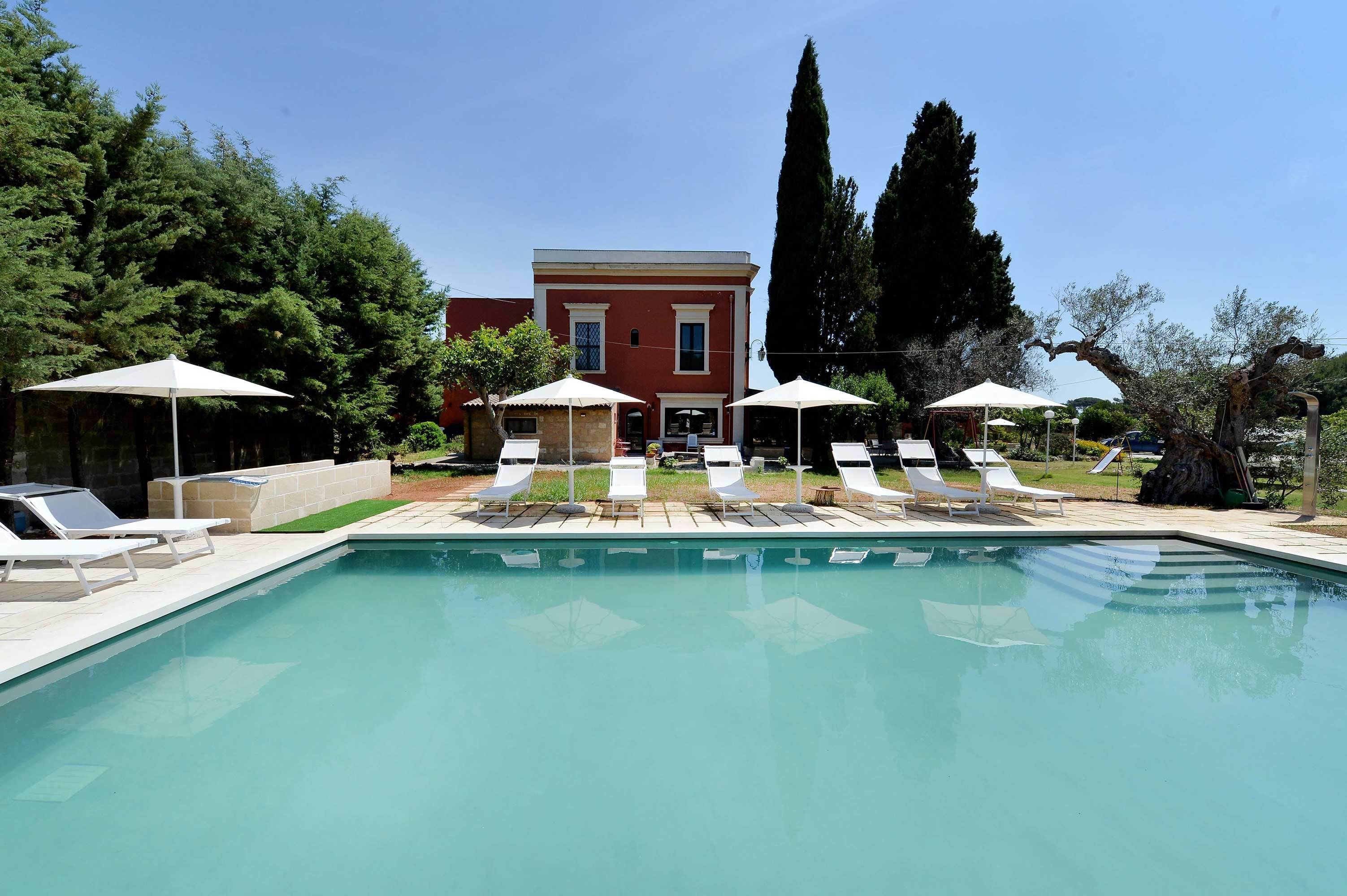 Una piscina con sillas de descanso y sombrillas, rodeada de árboles y un edificio.