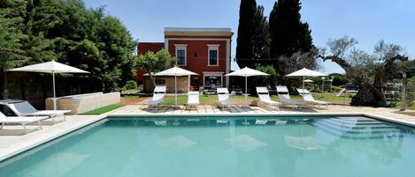 Una piscina con sillas de descanso y sombrillas, rodeada de árboles y un edificio.