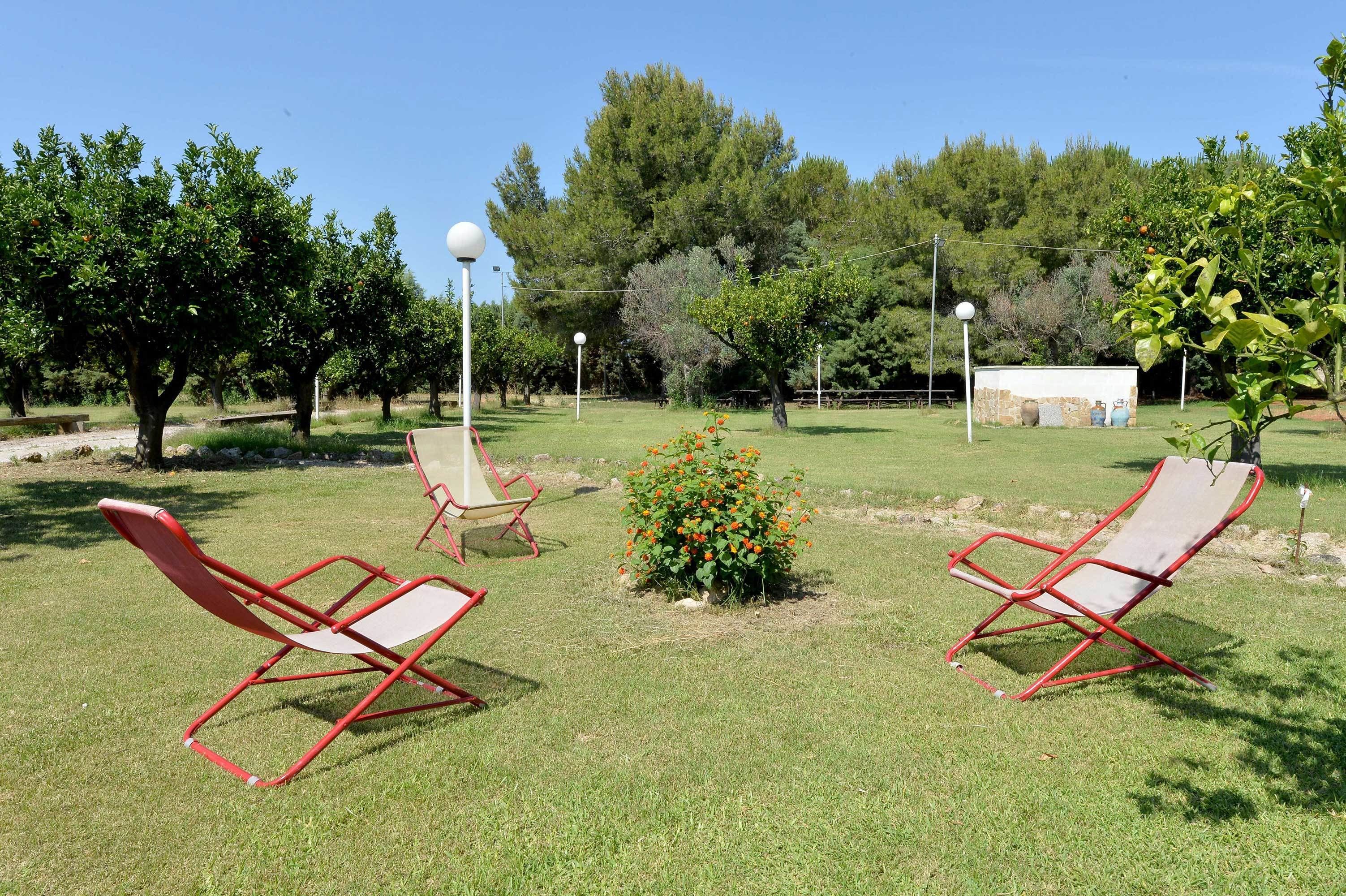 Un espacio al aire libre con dos sillones rojos, una pequeña zona de flores y un edificio blanco al fondo.