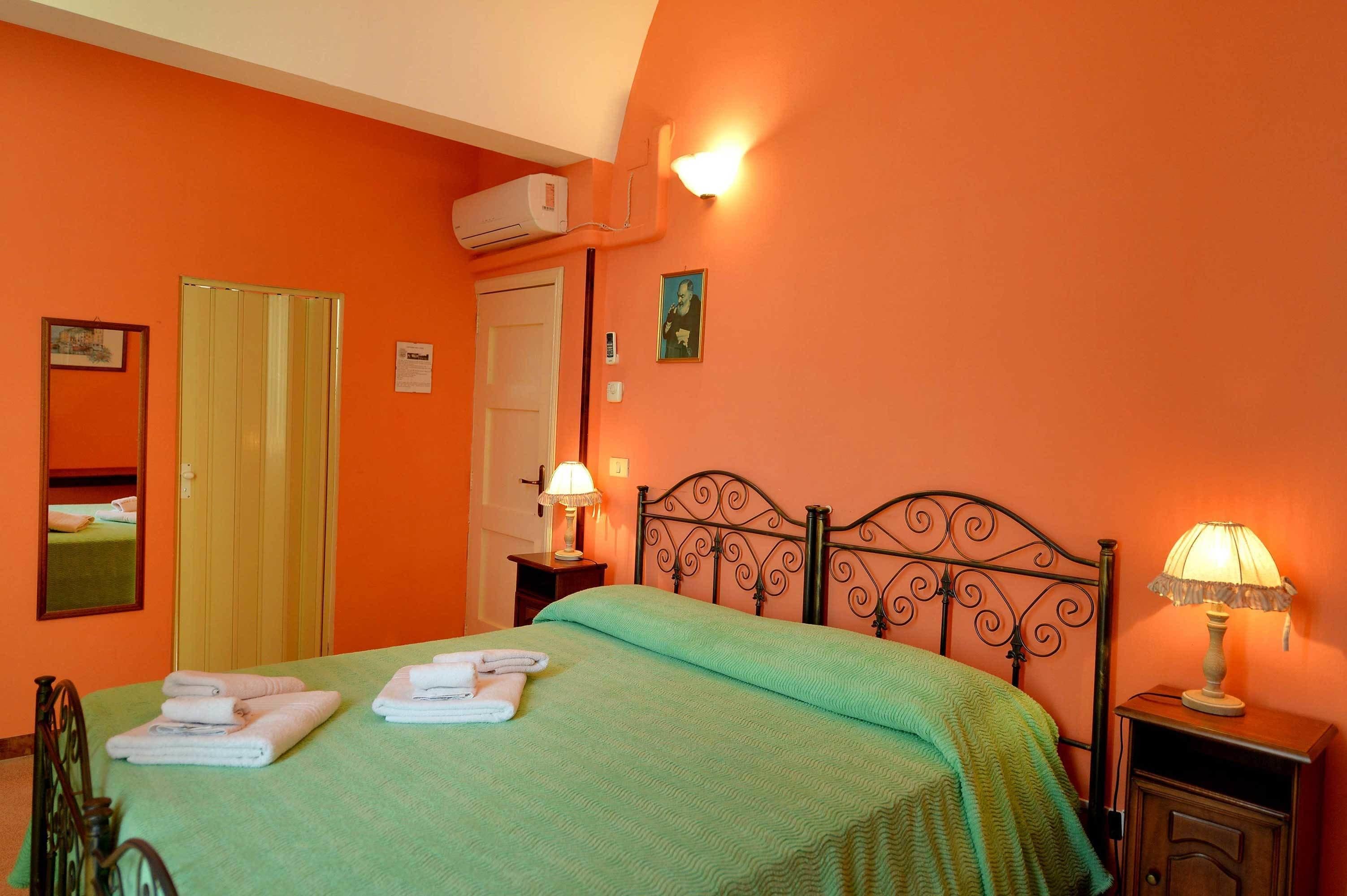 Una habitación con paredes anaranjadas, colcha verde, cabecero de metal negro, dos lámparas de noche y un espejo en la pared izquierda.