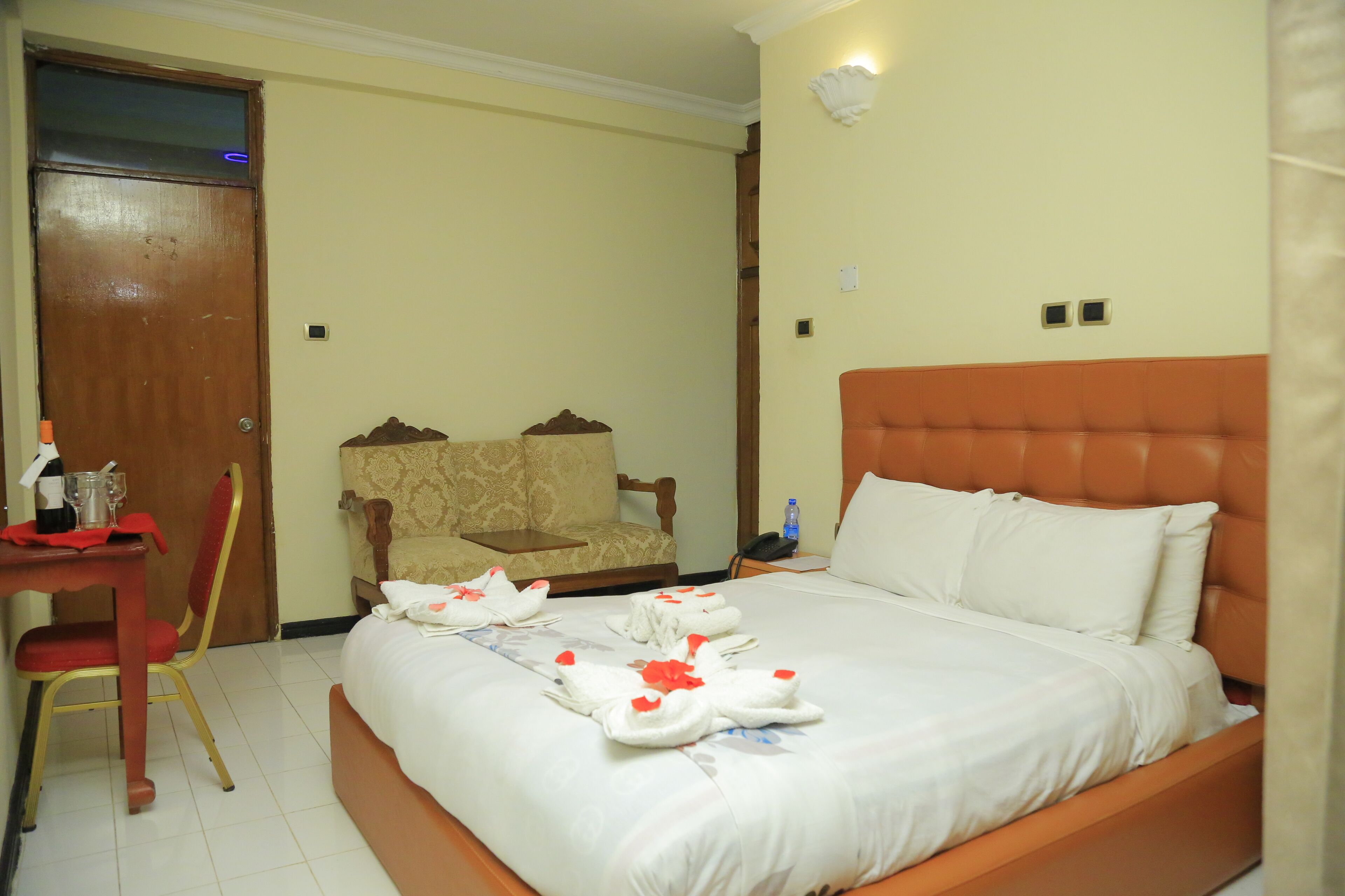 A hotel room with a bed, a chair, a table with a bottle, and a door.