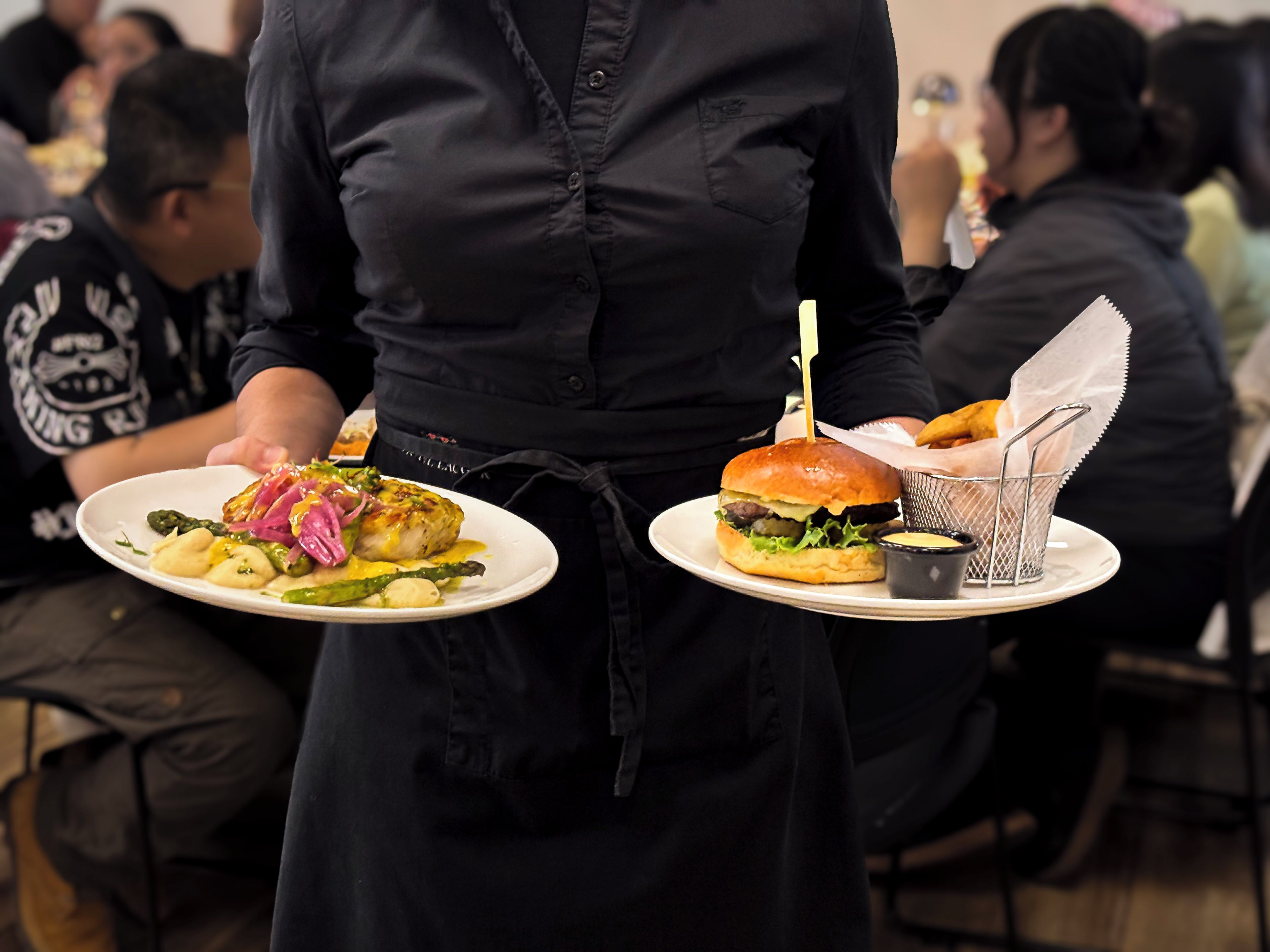 A waiter holding two plates of food, one with a burger and the other with a dish containing asparagus and other vegetables.