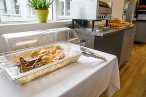 Een ontbijtbuffet met diverse broden en gebak, een Duolit koffiemachine, en een kamerplant bij het raam.