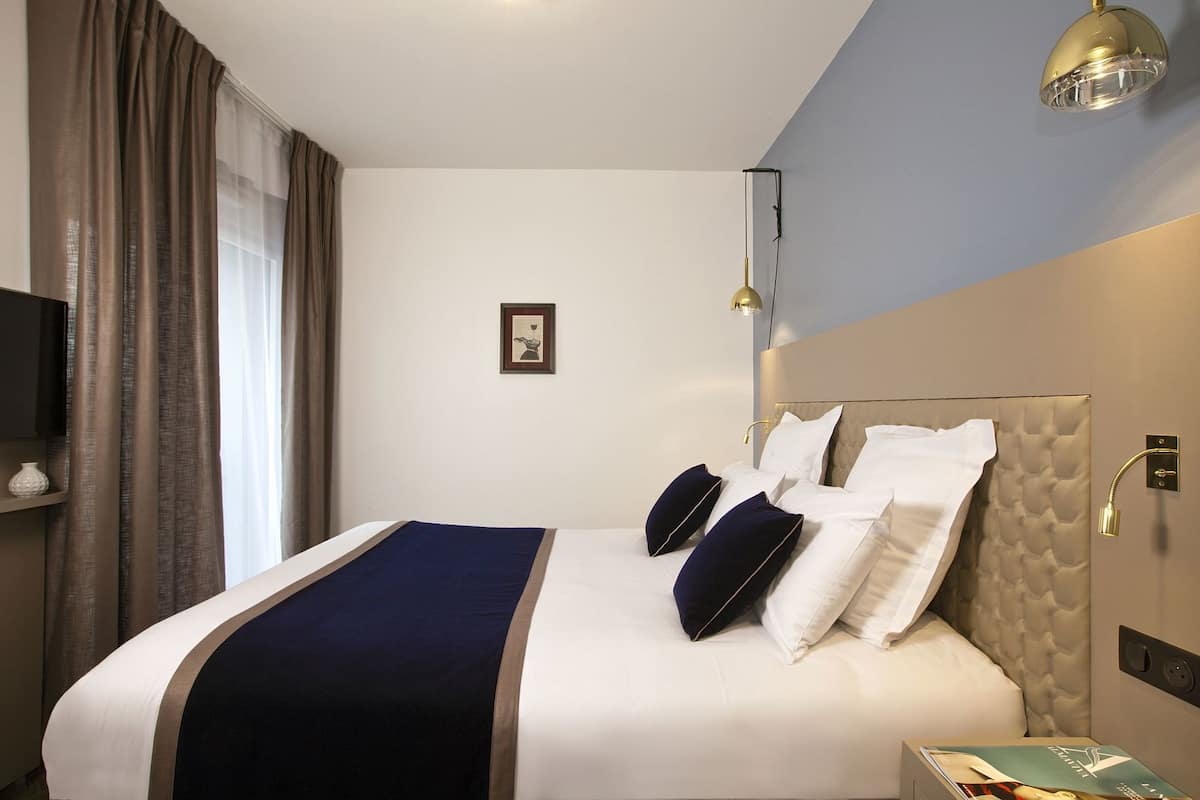 Habitación de hotel con una cama, una mesita de noche, un televisor montado en la pared y un ventanal con cortinas.