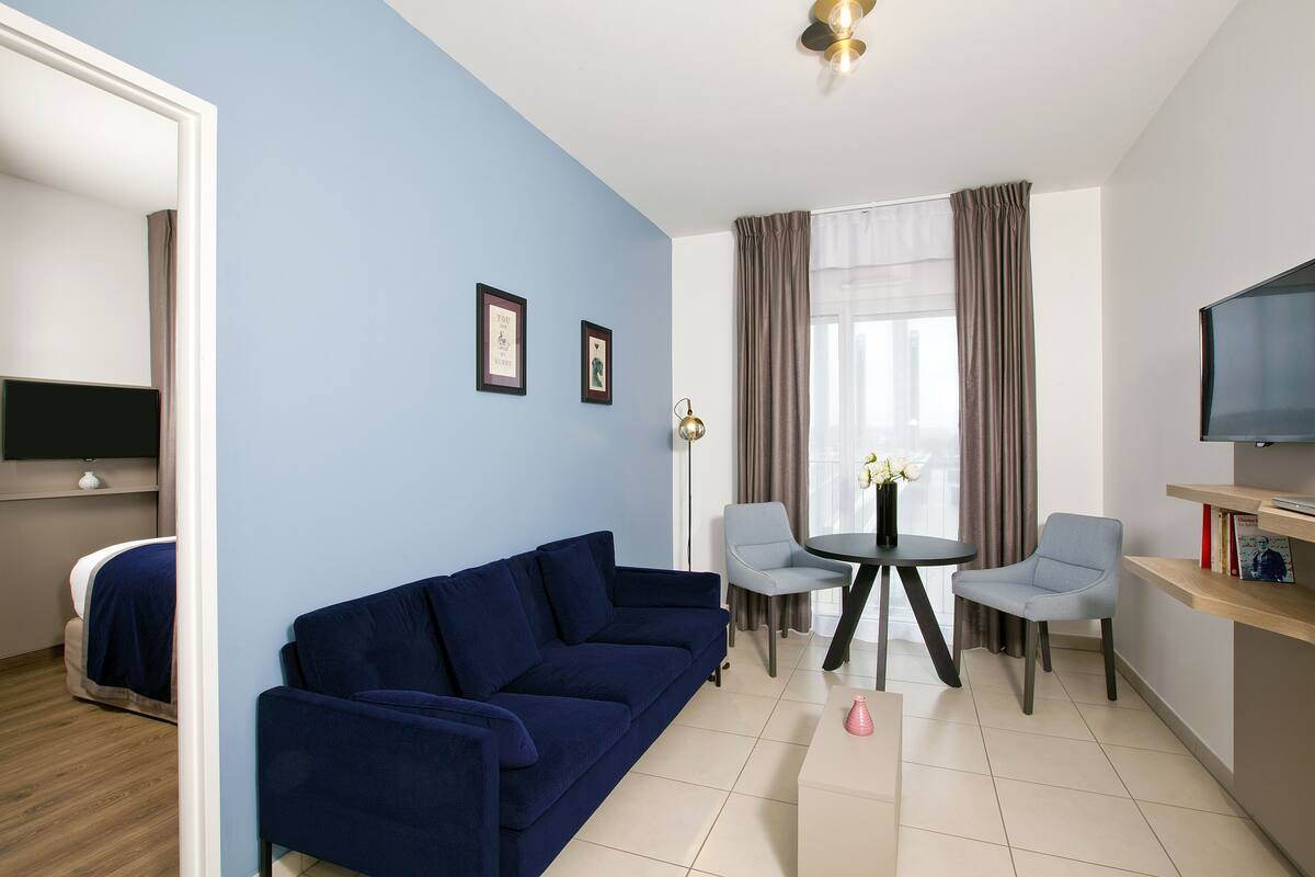 Una habitación moderna con pared azul, un sofá azul oscuro, dos sillas grises claras, una mesita redonda pequeña, un televisor sobre una estantería de madera y una cama con cabecera blanca.