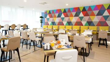 A modern dining area with multiple tables and chairs, a colorful geometric wall, and a plant in the corner.