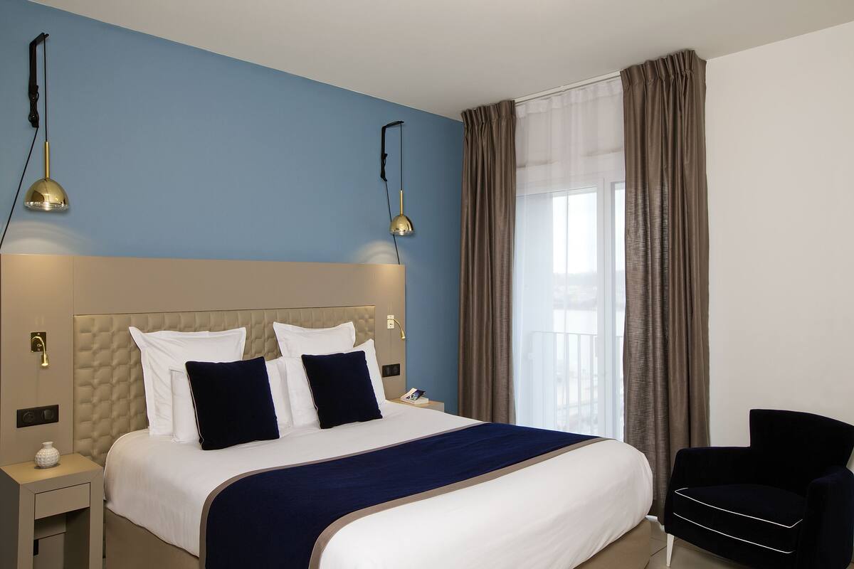 Una habitación de hotel con una cama grande, dos lámparas de noche, una mesita de noche, una silla y una ventana con cortinas.