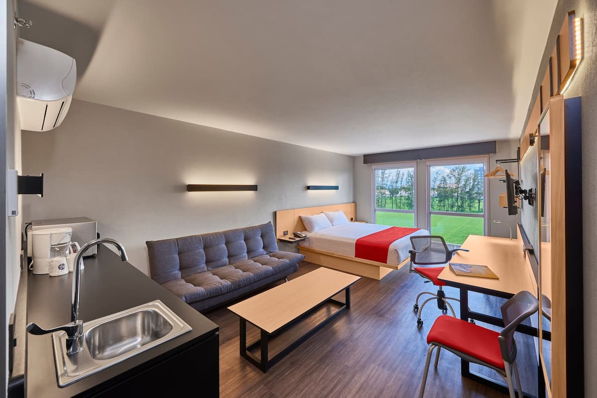 A modern hotel room with a kitchenette, a sofa, a bed, a desk, and a view of the outdoors.